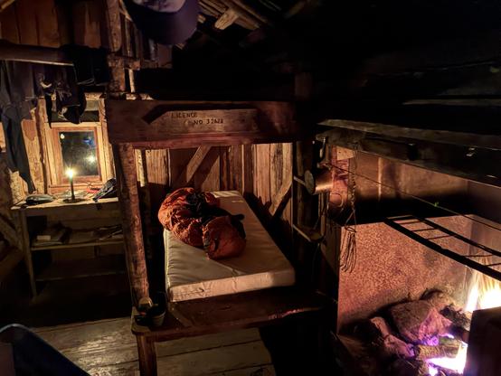 A firelit scene inside a very rustic goldminers hut - warm tones not reflecting the temperature inside the structure, furniture constructed from cut tree limbs, walls from slabs of sawn and adzed timber, open fire.