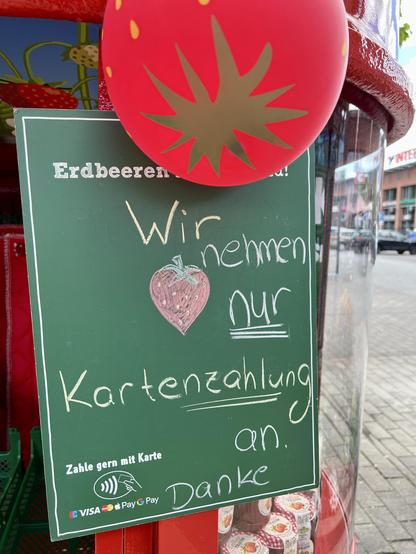 Ein grünes Schild an einem Obststand weist darauf hin, dass nur Kartenzahlungen akzeptiert werden. Auf dem Schild ist eine Erdbeere abgebildet und der Text „Wir nehmen nur Kartenzahlung an. Danke“ zu lesen.