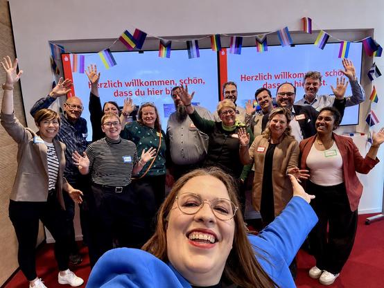 Ein Selfie auf dem 14 Menschen. zu sehen sind die fröhlich in die Kamera schauen. Im Hintergrund sind zwei von LGBTQIA+-Flaggen umrahmte große Monitore an der Wand zu sehen, auf denen steht "Herzlich willkommen, schön, dass du hier bist."