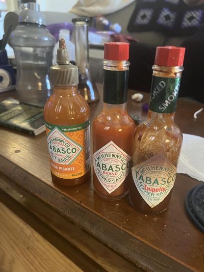 Three bottles of sauce are lined up on a wooden surface. The leftmost bottle is a large, translucent plastic squeeze bottle with a tan cap, containing an orange sauce, and features a label with the text "HENNY BRAND ABASCO SAUCE SALSA PICANTE", "PRESERVATIVE FREE", and "8 fl oz (236ml (236ml)". The middle bottle is a glass bottle with a red cap and black neck, containing an orange-red sauce, with a label reading "HENNY BRAND ABASCO PEPPER SAUCE" and "5 fl oz (148ml)". The rightmost bottle is a glass bottle with a red cap and a dark green neck label that says "SMOKED", "SMOKED", containing a thick, dark brown sauce, with a label reading "HENNY BRAND ABASCO CHIPOTLE SAUCE" and "5 fl oz (148ml)". The background is out of focus and contains a grey water bottle, a glass vessel, and a dark-colored couch.