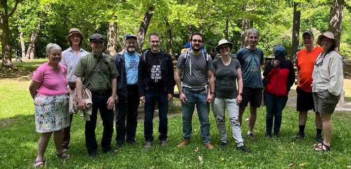 A group of white people (five women and six men) standing side by side in a lush green park, of various ages, looking like they have have been coerced into a group photo. One of them is in orange and looks like a muppet that was shaved.