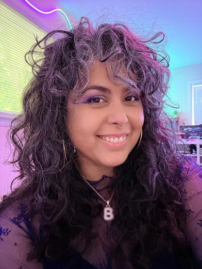A woman with black and gray wavy hair smiling into the camera with colorful lighting behind her. She's also wearing a pendant with the letter "B" and wearing a floral top.