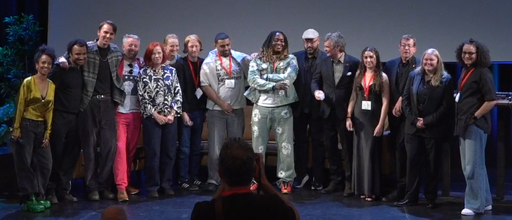 A group of roughly fifteen conference speakers and organizers posing together on stage for a photo, with an audience member's silhouette visible in the foreground. Several people are wearing red lanyards with name badges.