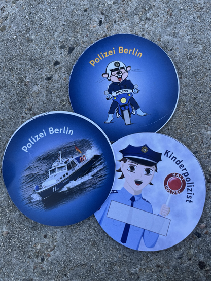Drei runde Sticker der Polizei Berlin, eines mit einem Foto eines Boots, eines mit einer mangaartigen Figur mit Polizeikelle in der Hand, und eines mit einer gezeichneten Maus in Uniform auf einem Motorrad