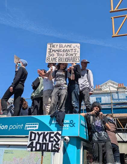 A young person in a crowd stood up on top of a kiosk holds a sign that says they blame immigrants so you don't blame billionaires. 
Down below is a sign that reads Dykes 4 Dolls